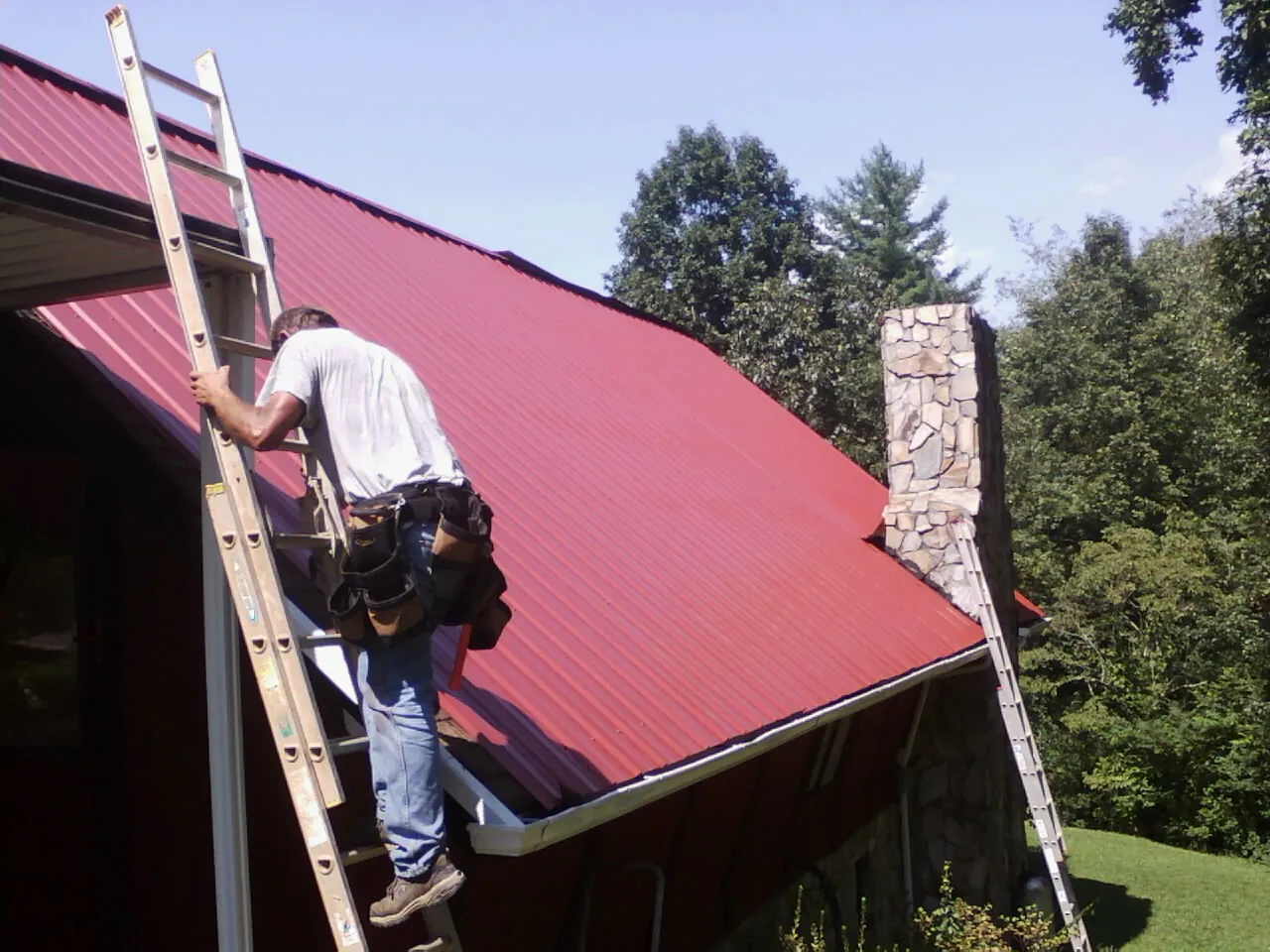 Roofer on a ladder beside a red metal roof and stone chimney — Soffit & Fascia Repair by Roofing Brothers in Onley